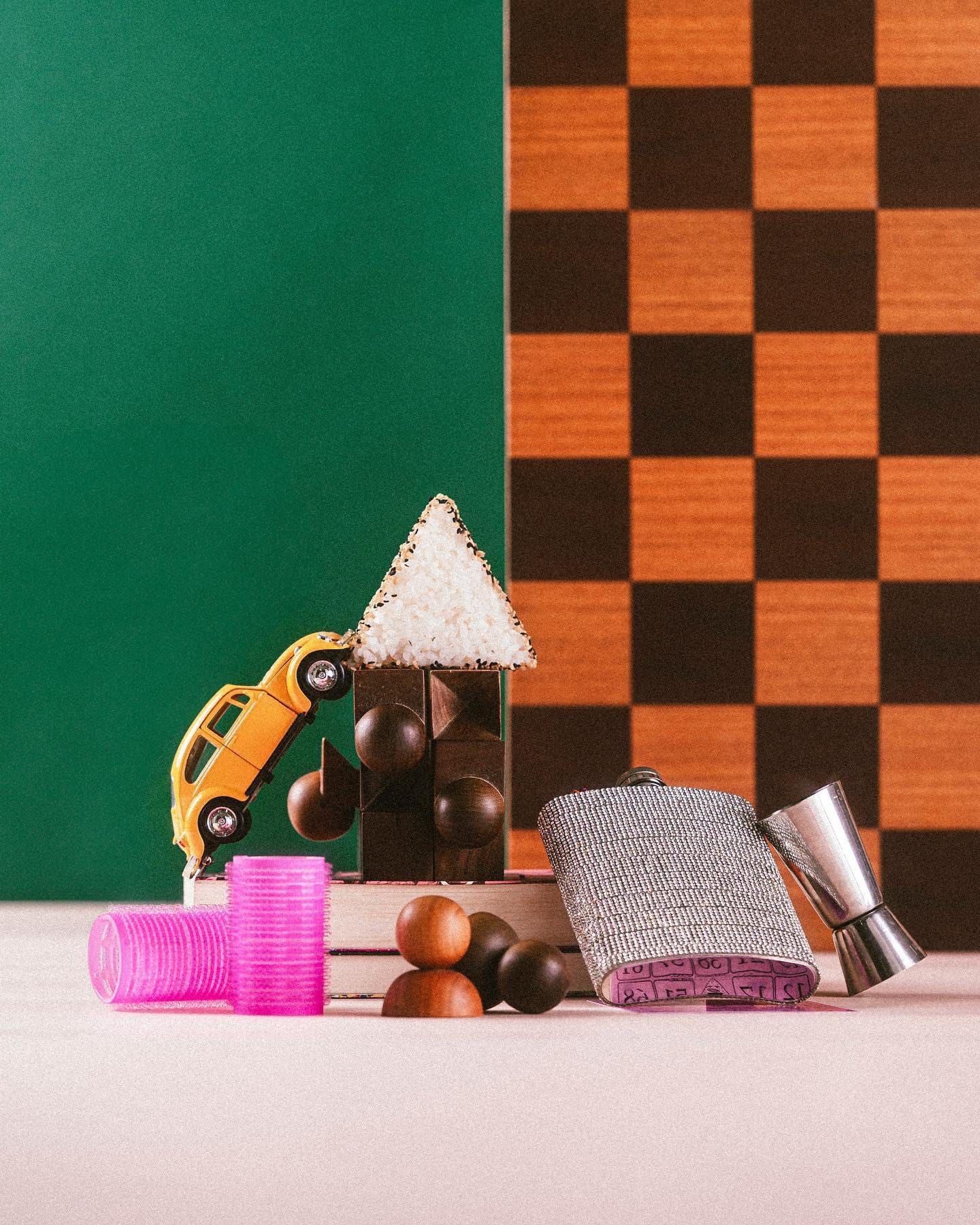 Still: onigiri sobre uma torre de objetos, com um carrinho amarelo, bobes e um cantil, diante de um tabuleiro de xadrez