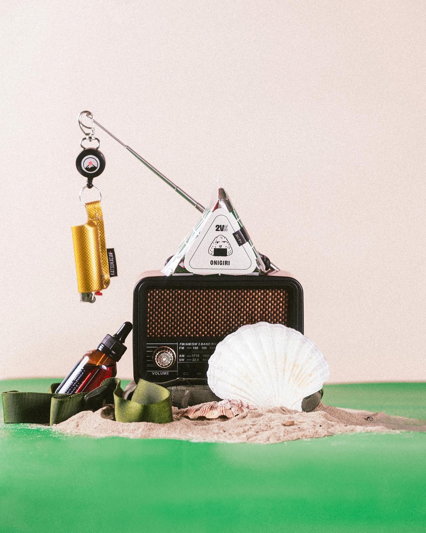 Still: onigiri embalado pendurado sobre um rádio antigo, uma concha e areia