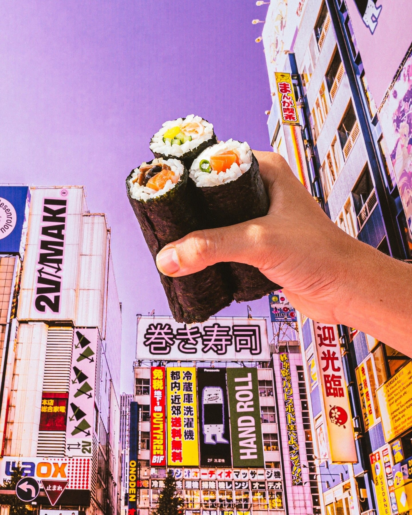 Mão segurando três hand rolls diante de prédios com letreiros em japonês e um painel 2V Maki
